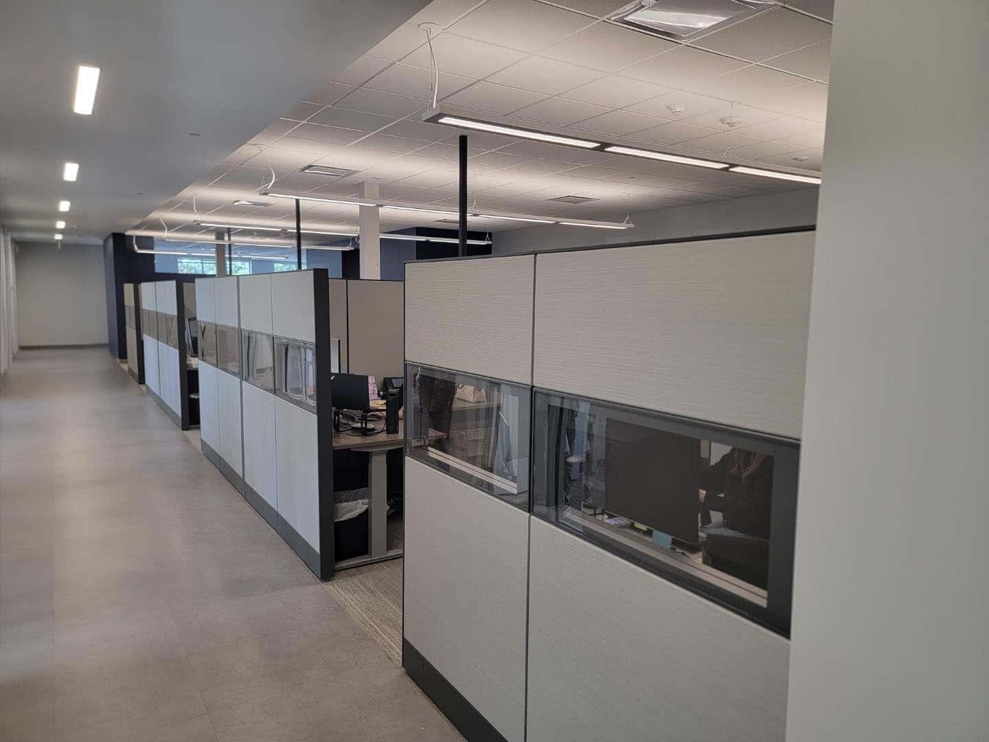 Row of gray cubicle workstations with glass-topped privacy panels along an office hallway
