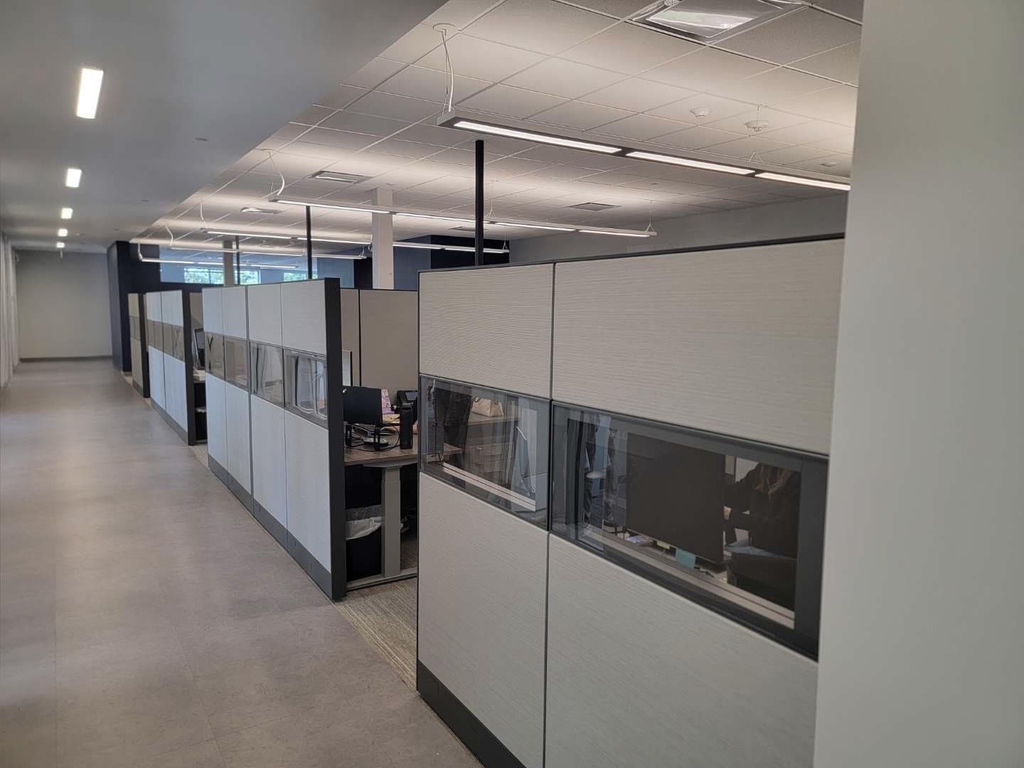 Row of gray cubicle workstations with glass-topped privacy panels along an office hallway